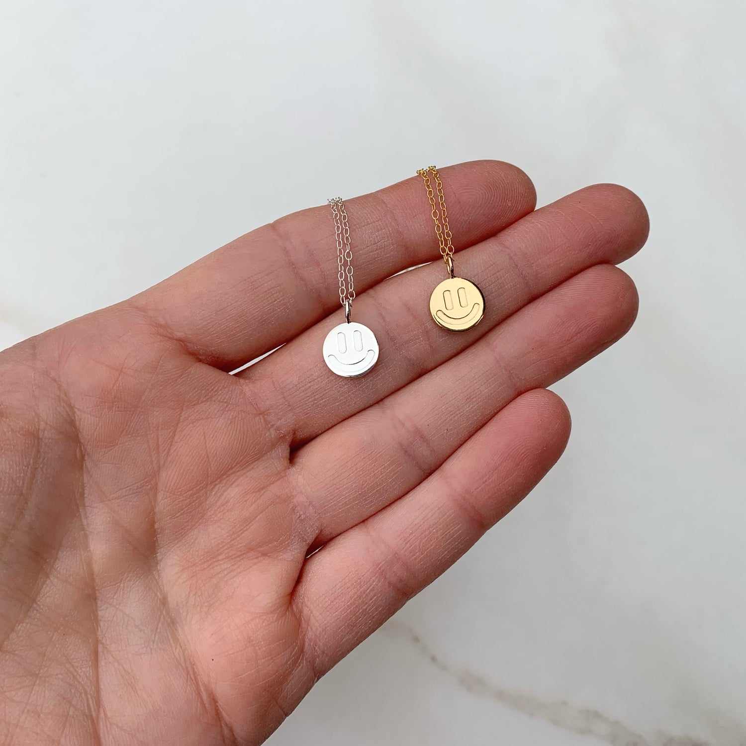 a sterling silver and a gold smiley face necklace being held in a hand over a white background