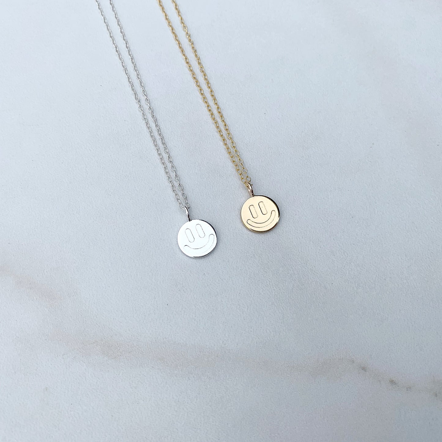 one sterling silver smiley face necklace next to a gold smiley face neckalce on a white background