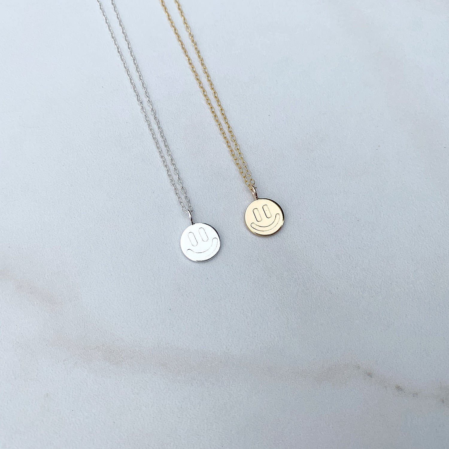one sterling silver smiley face necklace next to a gold smiley face neckalce on a white background